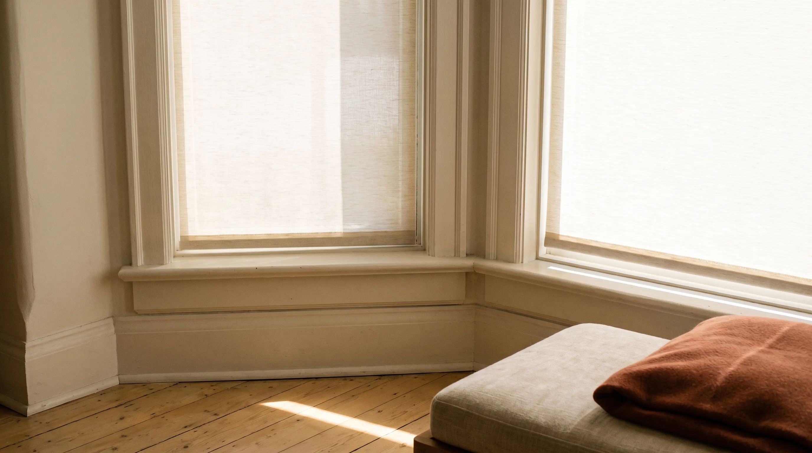 The corner of a bay window where two roller blinds meet at a painted mullion, each fitted inside its own reveal with a narrow shadow line between them and daylight coming through both fabrics