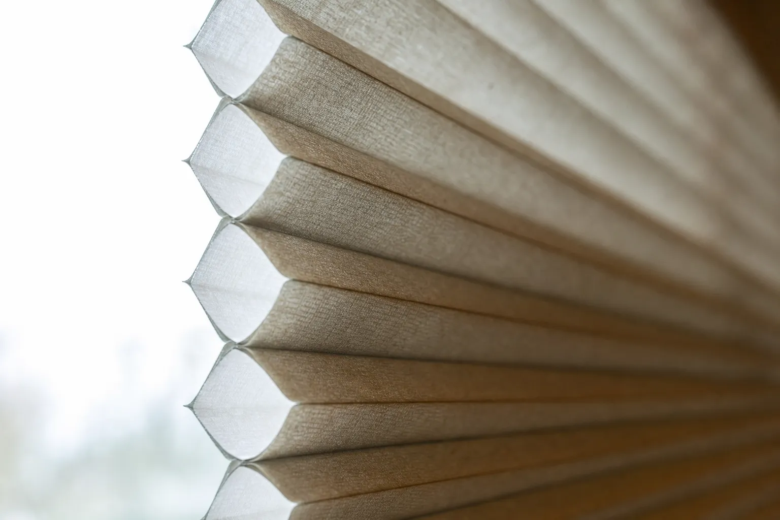 A close side-on view of a honeycomb shade against a bright window, the light passing through so the hollow six-sided cells and the woven texture of the fabric are visible in cross section