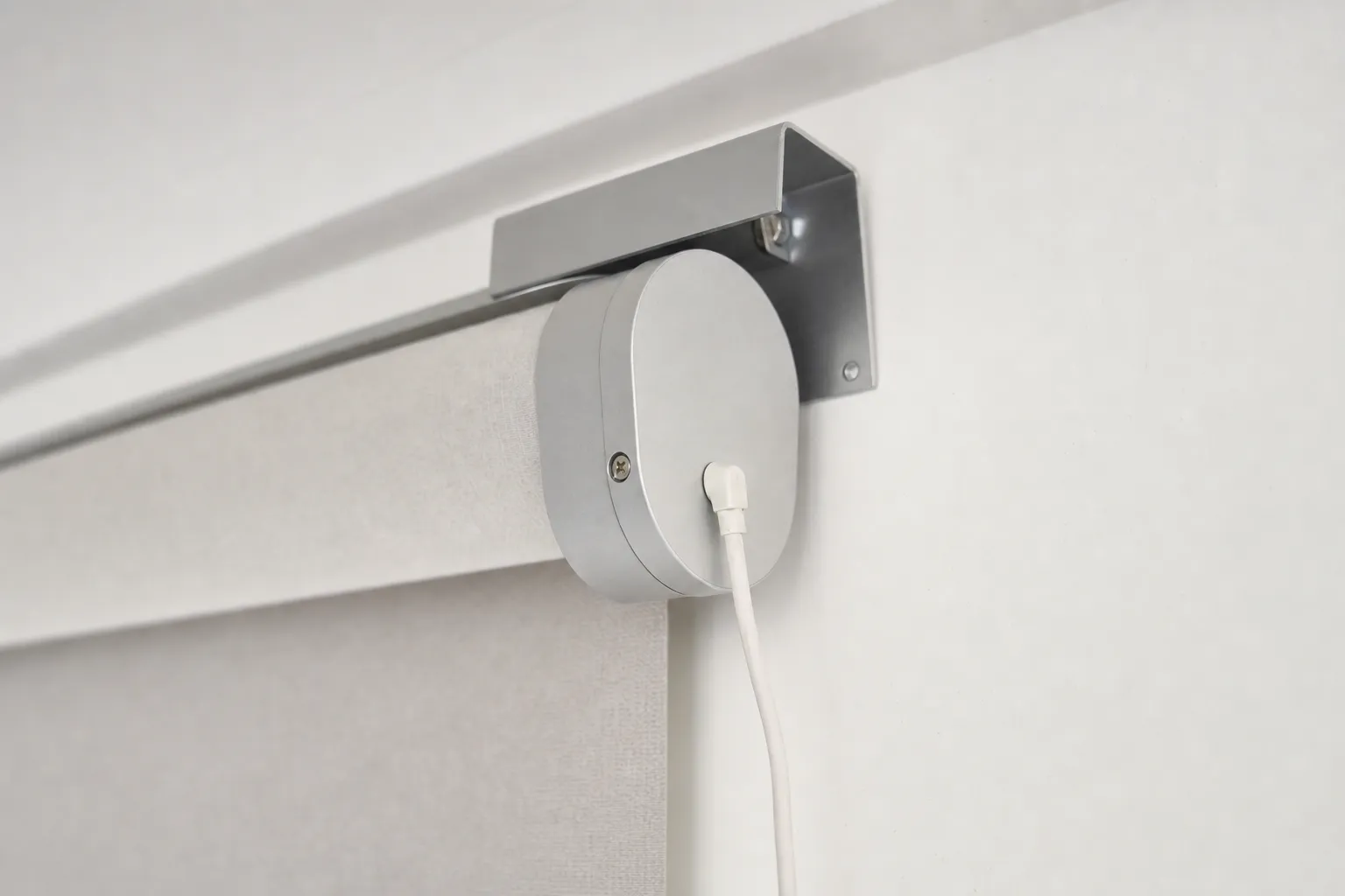 A close view of the end of a roller blind where a rounded aluminium end cap sits under its bracket, a slim white charging cable plugged into the cap and hanging down the wall