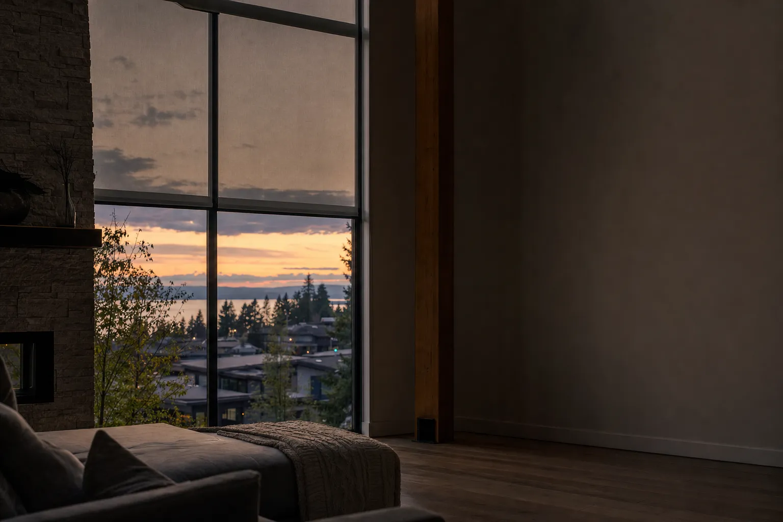 A dusk room with a two-storey window, a screen blind lowered across the upper section while the lower glass stays clear over a lit town and a sunset, and a large unlit wall to the right
