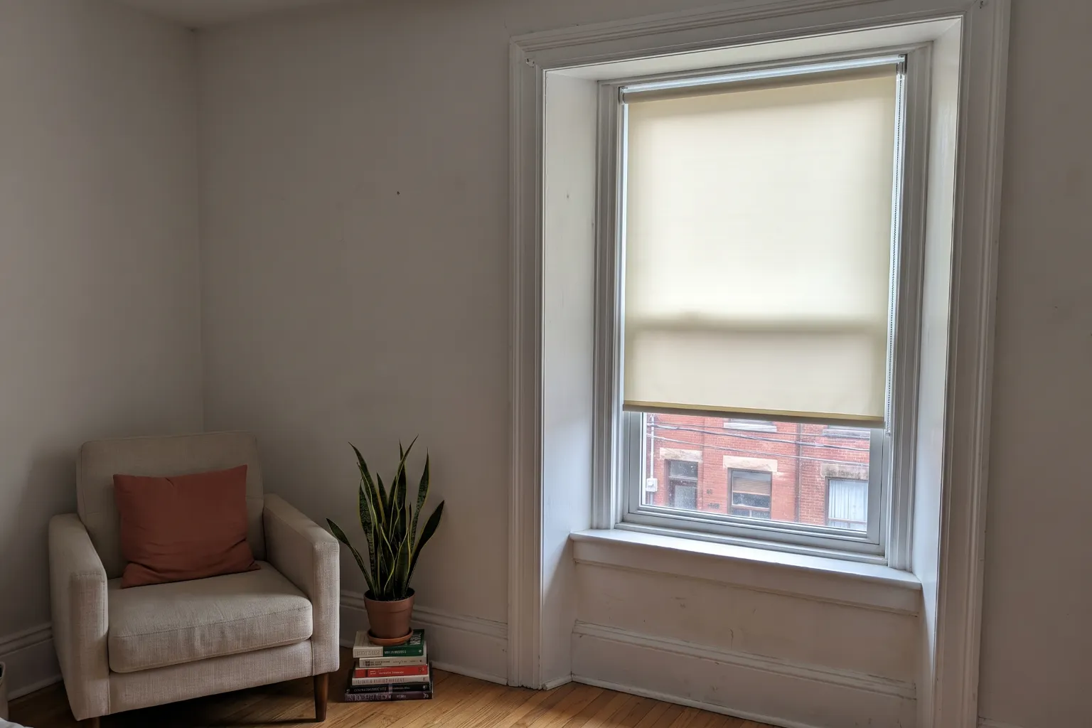 A cream roller blind lowered two thirds over a tall window in the deep painted reveal of an older Toronto house, its roller tension-fitted inside the recess so the round end caps press flat against the reveal with no brackets, screws or drill holes in the untouched white trim, a slim control chain down the right side, red-brick houses across the street through the clear glass below, and a linen armchair with a terracotta cushion, a snake plant and a stack of books to the left of the window