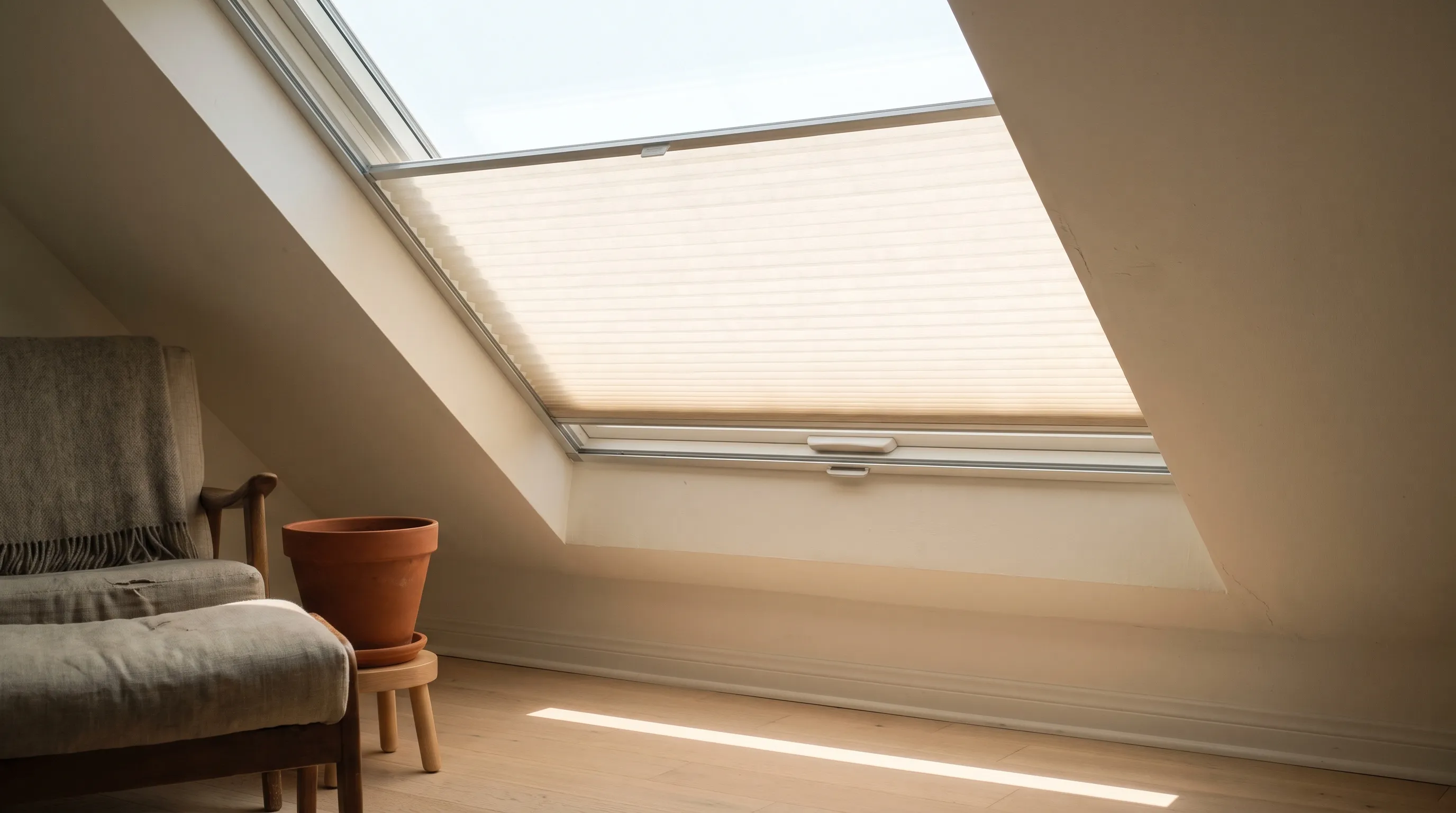 A close view of a pleated skylight blind on a sloping ceiling, the warm-white fabric held taut in slim aluminium side channels with a slim bottom bar, and bright sky through the glass above it